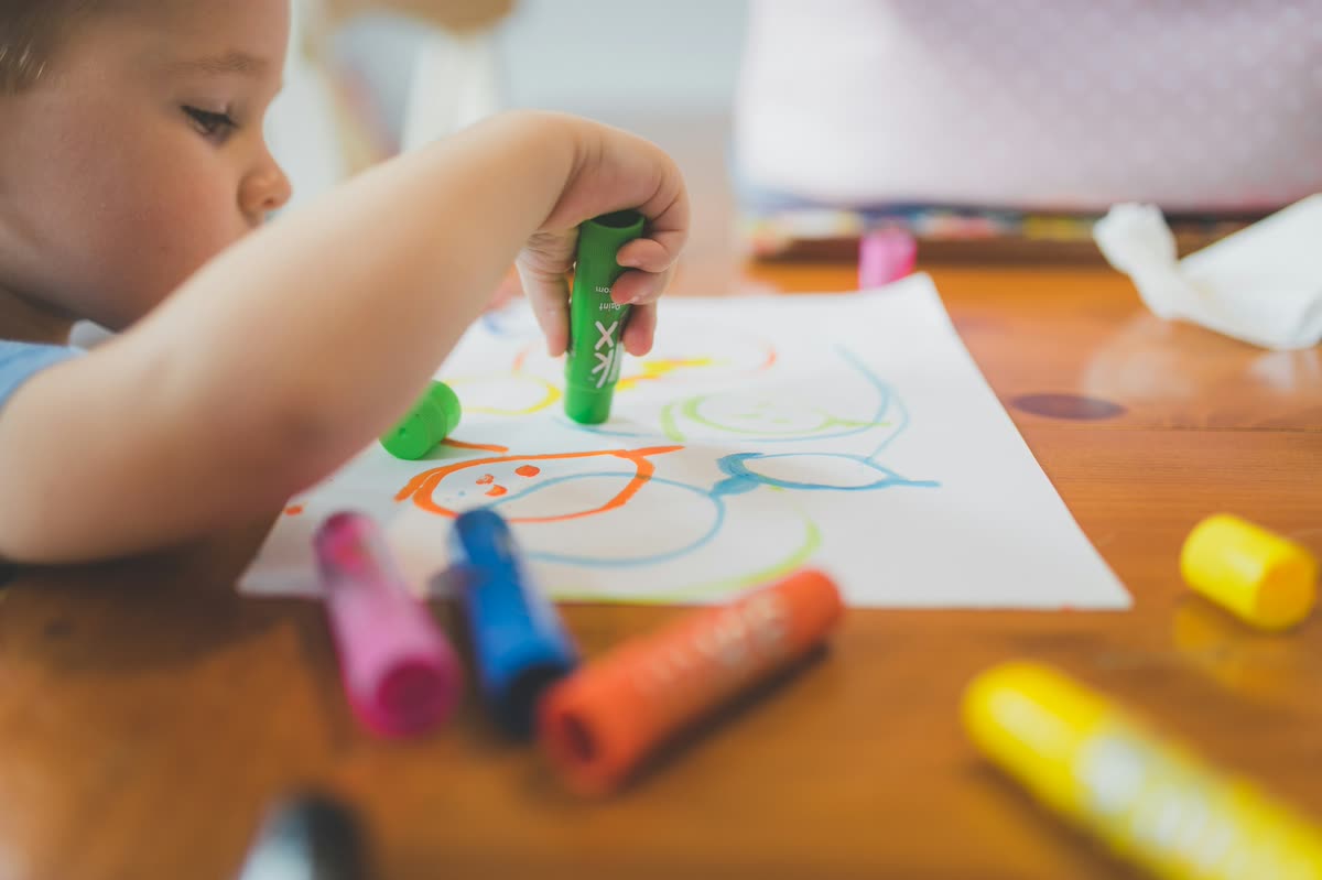 A young child colors at the kitchen table with crayons scattered across the paper